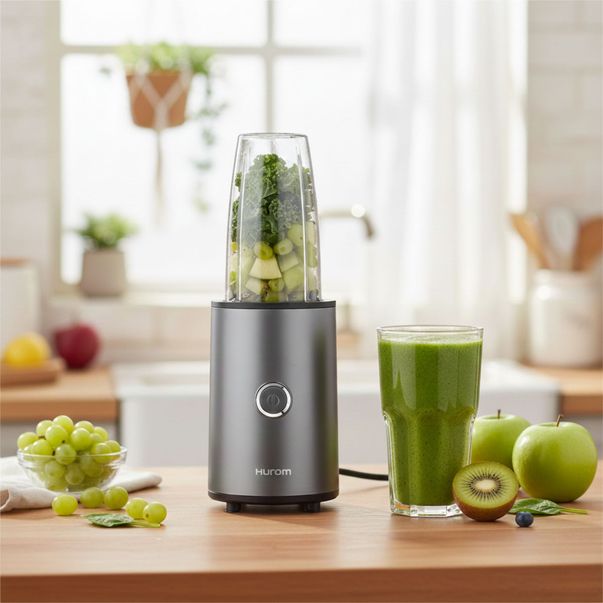 Hurom Power Blender mit gefülltem Behälter im Küchenambiente mit einem Glas Grünen Smoothie und Früchten
