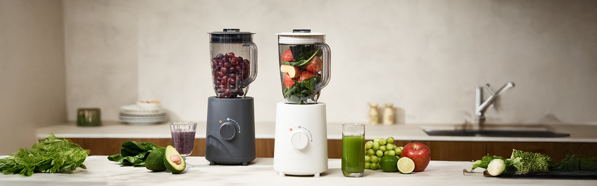 Hurom Standmixer in Milestone Gray und Mushroom befüllt mit Zutaten auf Küchenanrichte mit Obst und Gemüse